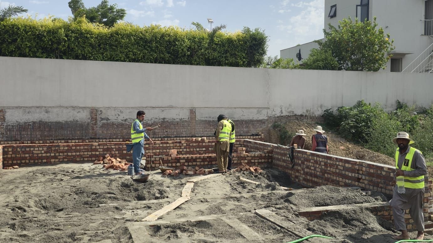 Workers building brick plinth walls beside a grey boundary wall at a house construction site in Lake City Lahore